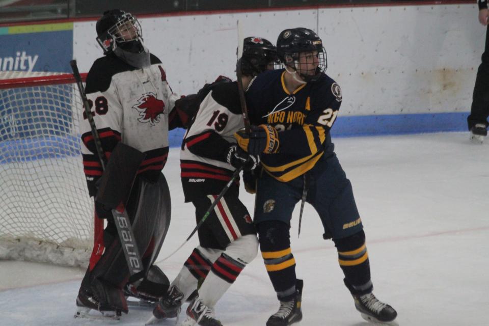 Devils Lake forward/defenseman Carson Hanson (middle, white) attempts to block off a screen set by Fargo North forward John Martin (right, blue). Devils Lake goaltender Zach White (left, white) watches for the puck.