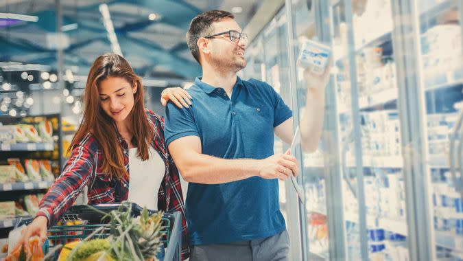 A young couple pushing a shopping cart filled with produce, picking up some cheese from the fridge in the dairy aisle at the supermarket.