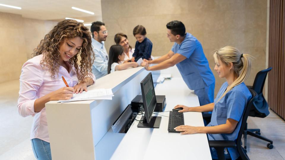 Beautiful latin american patient filling in a form at the hospital's front desk all smiling - Incidental people at background.