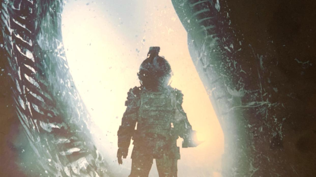  A figure in a spacesuit stands in an opening, leveling a torch toward the foreground. 