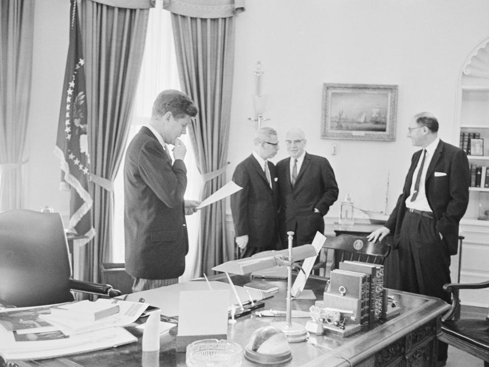John F. Kennedy, accompanied by Labor Secretary Arthur Goldberg, Commerce Secretary Luther Hodges, and Myer Feldman, Associate Counsel, scans some papers in the Oval Office.