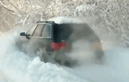 A black SUV drives through heavy snow on a forest road, snow billowing around the vehicle as it moves