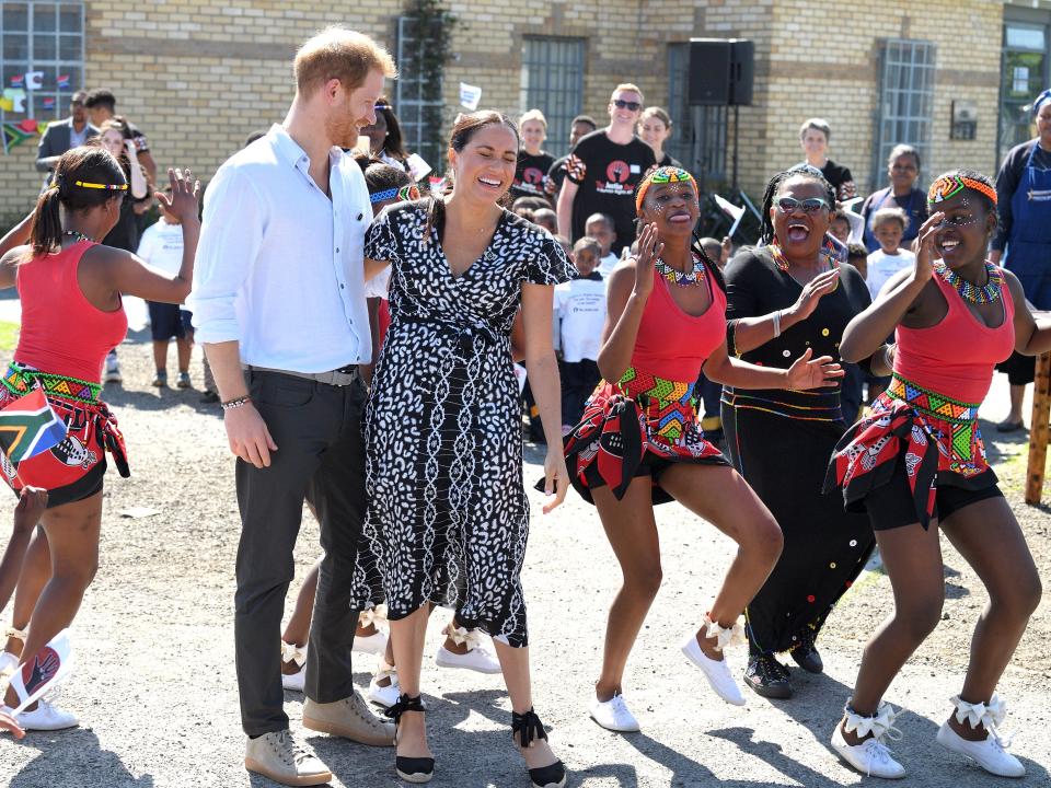 Meghan and Harry embracing and laughing while women dance around them outside.