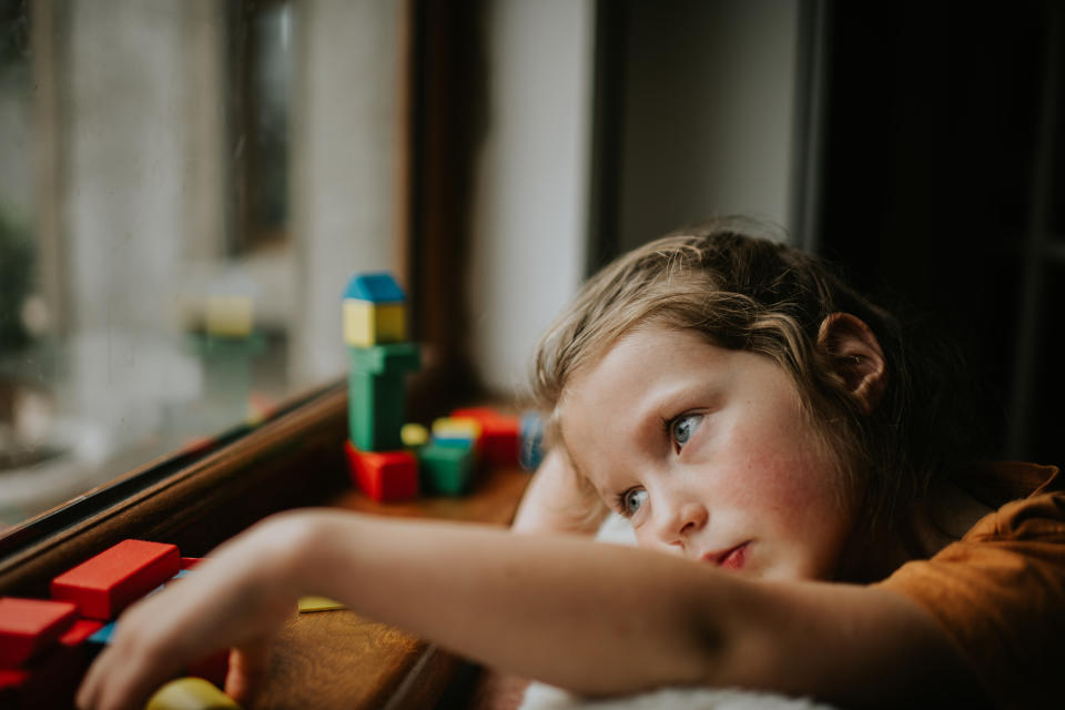 A young child looks bored as she half heartedly stacks her coloured wooden building blocks beside a window. Space for copy.