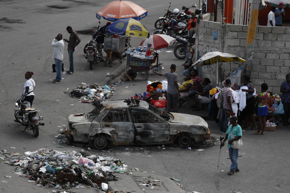 Vendors sell from sidewalk stands and people wait for public transport, alongside a car burned during Monday protests, in the Delmas district of Port-au-Prince, Haiti, Wednesday, Oct. 2, 2019. There was a relative pause in disturbances that have wracked Haiti’s capital for weeks as protesters have tried to drive President Jovenel Moïse from power. (AP Photo/Rebecca Blackwell)