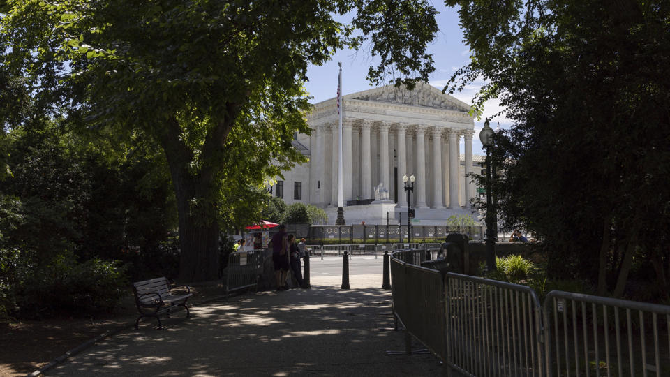 View of Supreme Court building through a clearing of trees, beyond a park bench and sidewalk vendor.