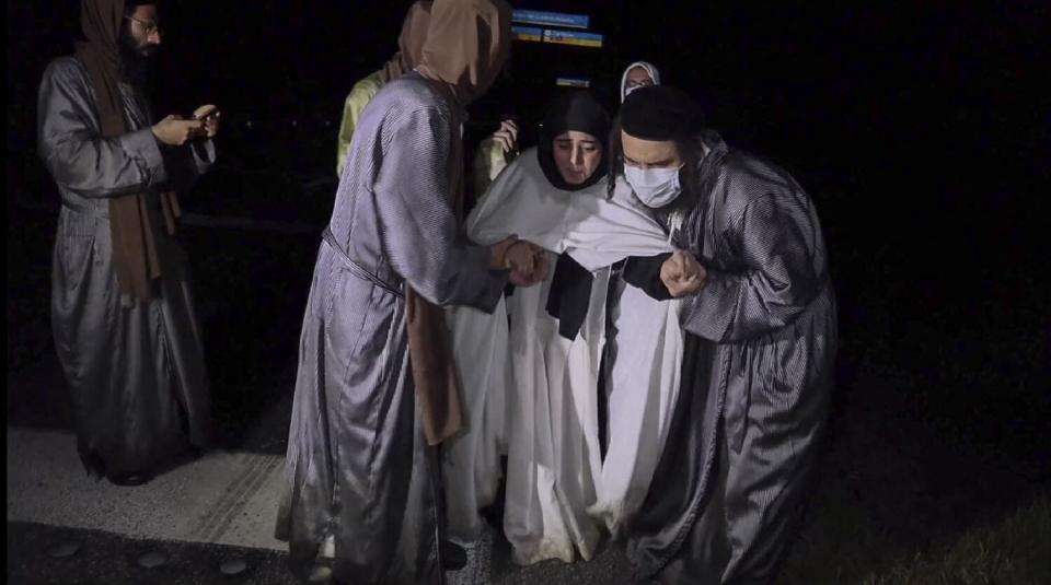 People in gray robes assist a person in a white robe and dark head covering as a man stands nearby on the left