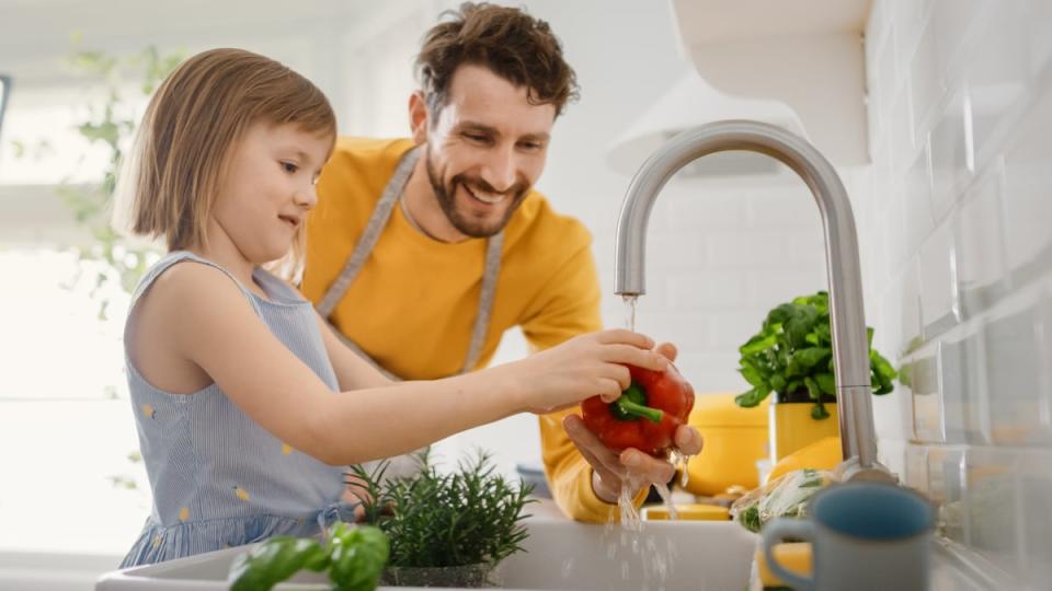 An adult smiles while helping a child rinse a bell pepper at a sink.