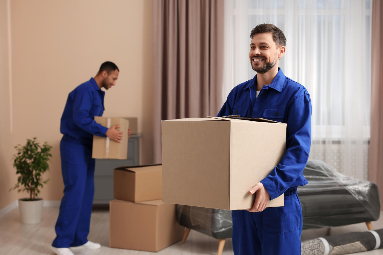 Two male movers are pictured moving cardboard boxes in a new house