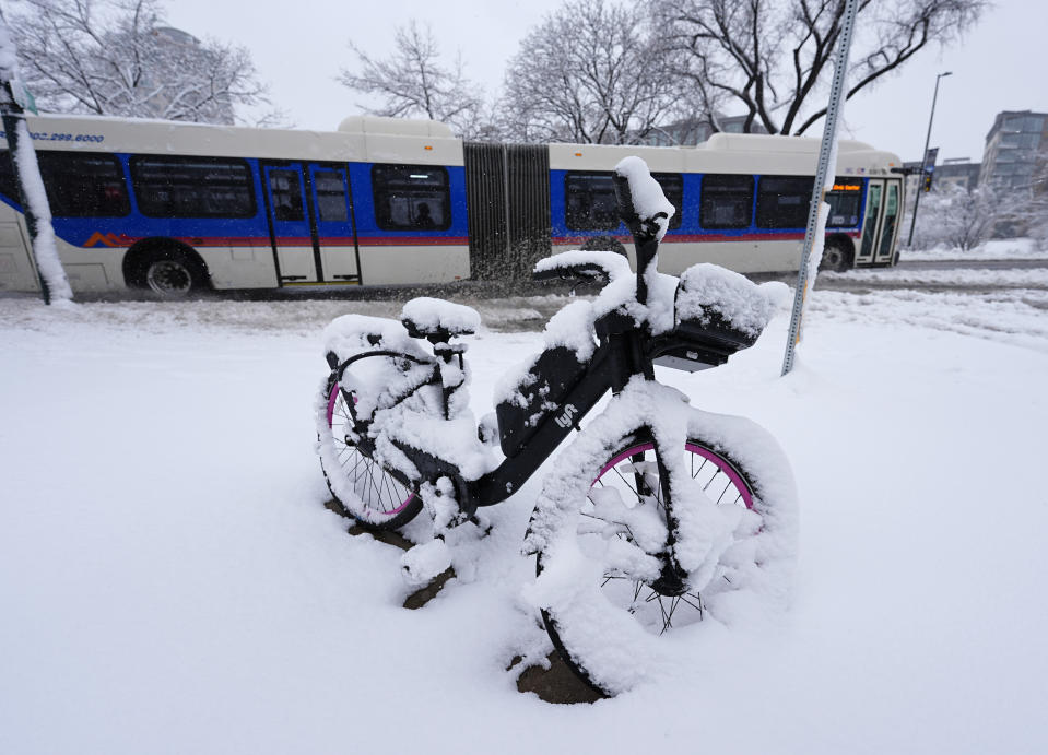 A bicycle sits covered with snow as a city bus moves along the westbound lanes of Speer Boulevard as a late winter storm dropped up to a foot of snow Thursday, March 14, 2024, in Denver. Forecasters predict that the storm will persist until early Friday, snarling traffic along Colorado's Front Range communities. (AP Photo/David Zalubowski)