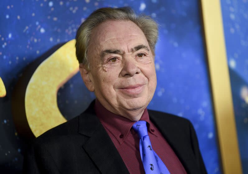 Andrew Lloyd Webber smiling in a black suit, maroon collared shirt and purple tie against a blue and gold background