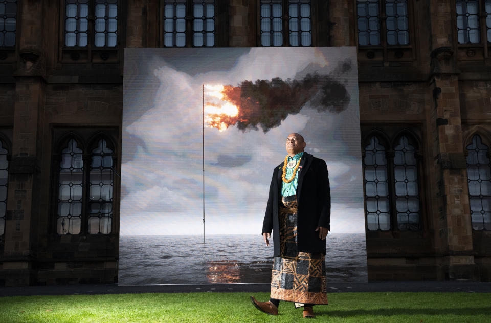 Tongan activist Uili Lousi stands alongside 'Flare Oceania 2021', created by artist John Gerrard, a real-time moving image showing a simulation of the seas around Tonga with the flag/flare embedded in it, displayed on the South Facade of the University of Glasgow to mark Ocean Day, at the COP26 climate summit, in Glasgow, Scotland, Friday, Nov. 5, 2021. (Jane Barlow/PA via AP)