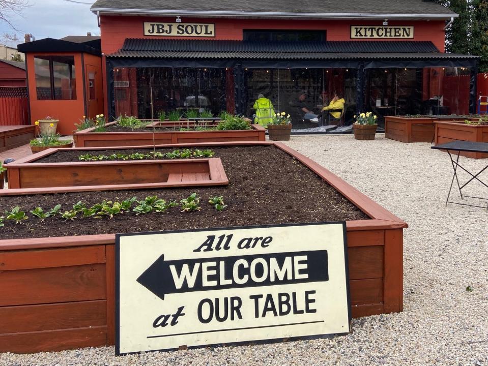 The outside of the red and black restaurant and front garden with wooden planters and plants. A sign leans against a planter and reads "all are welcome at our table."