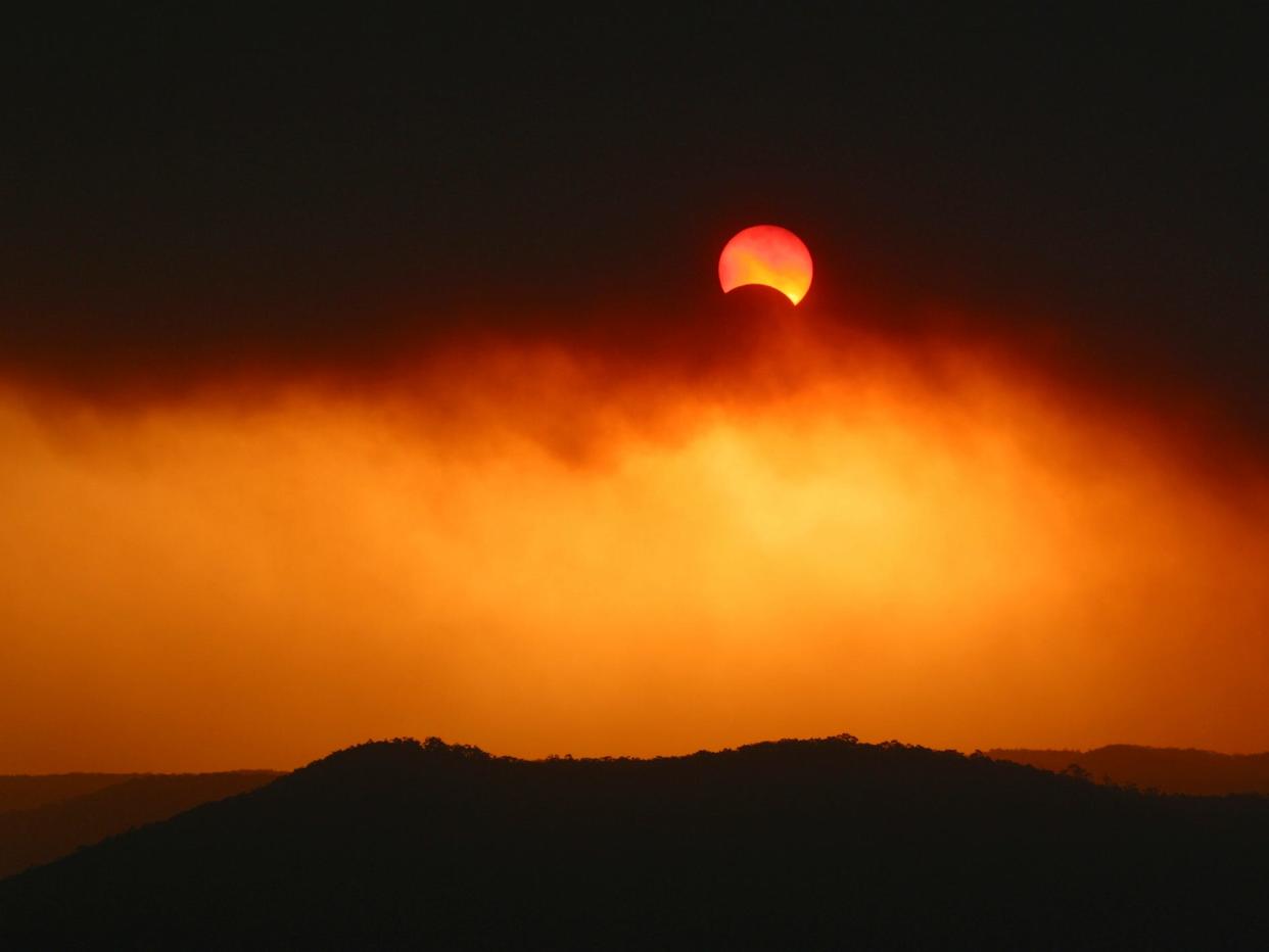 A partial solar eclipse appears fire red through thick plums of bushfire smoke.