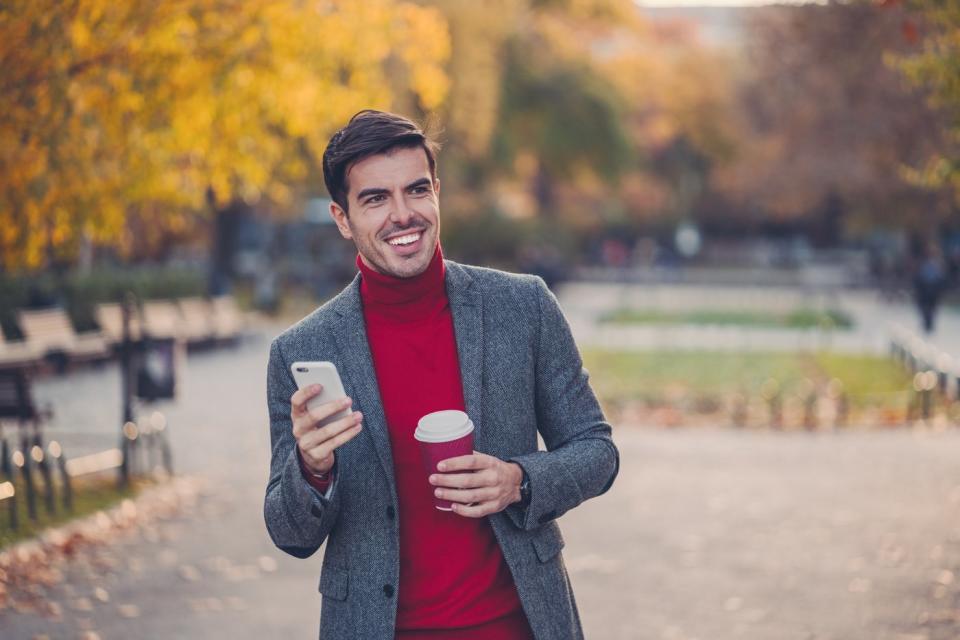 A person walks through a park in the fall holding a phone and a cup of coffee.