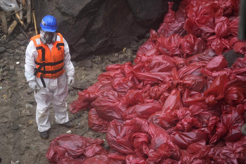 A worker stands next a red bags filled with oil waste during a clean-up campaign on Cavero Beach in the Ventanilla district of Callao, Peru, Tuesday, Feb. 15, 2022. One month later, workers continue the clean-up of beaches after contamination by a Repsol oil spill. (AP Photo/Martin Mejia)