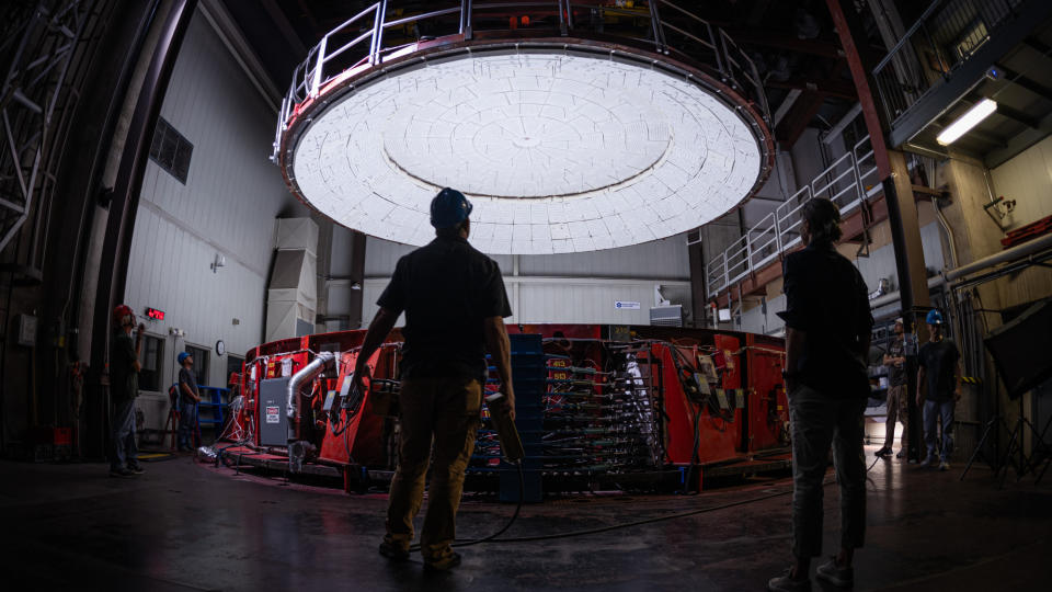 The lid closes on a large red furnace inside a white-walled building while two people watch.