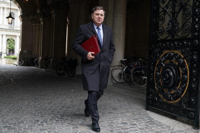 Mel Stride, in a long grey coat, walks through some black ornate gates on a cobbled path. He is carrying a red folder under one arm.