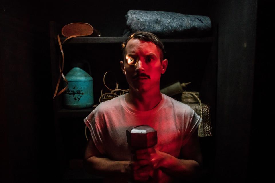 A mustachioed man stands in fear in a small closet while holding a dumbbell