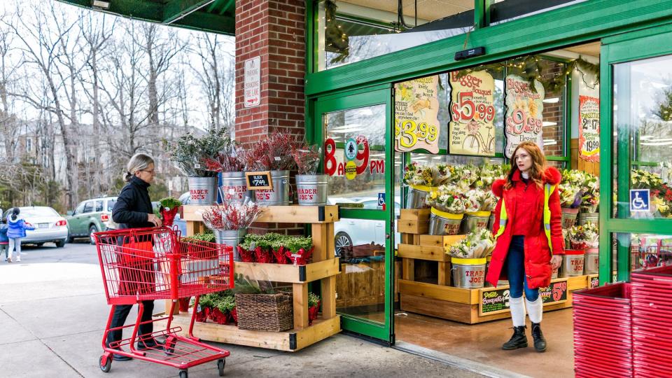 Trader Joe's employee in red clothes with customer, trolley shopping carts by store entrance doors outside women, winter flower pots, gardening plants in Virginia