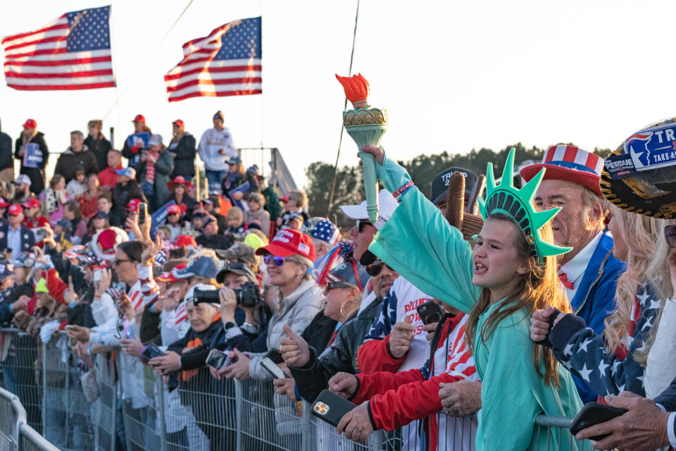 People at a rally, many of whom wear American-flag printed hats and jackets, along with a child wearing a Statue of Liberty costume, stand behind a low fence near two American flags against a white sky.