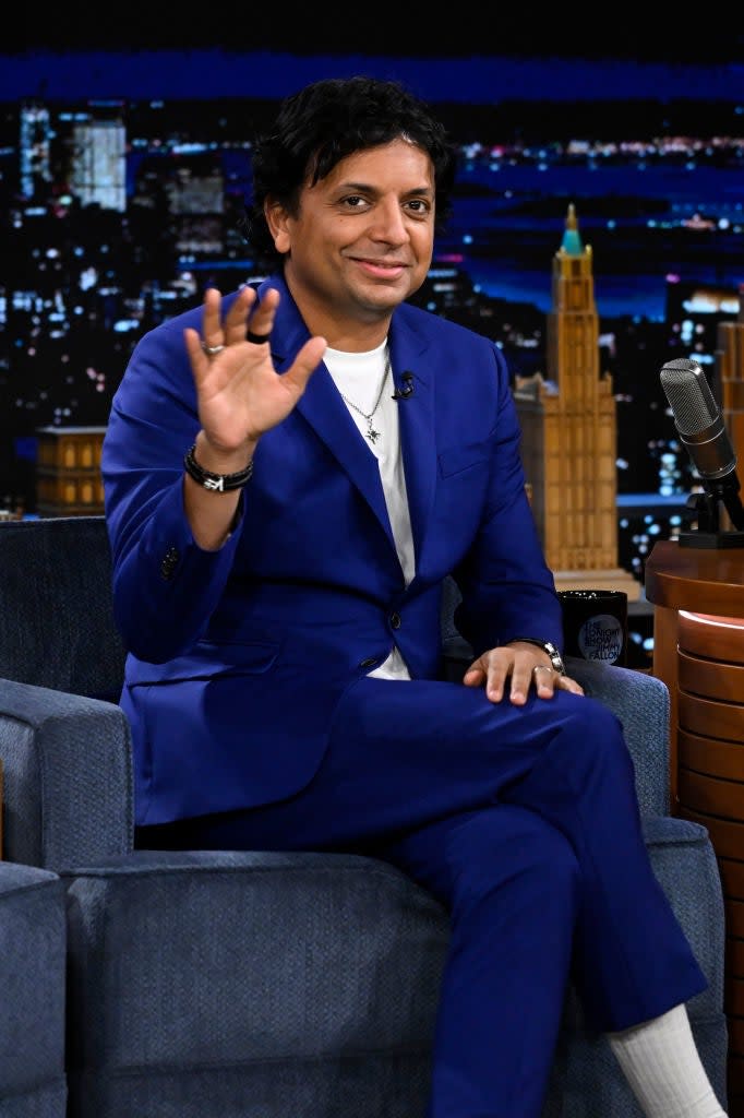 Man in a blue suit sitting on a talk show set, waving to the camera