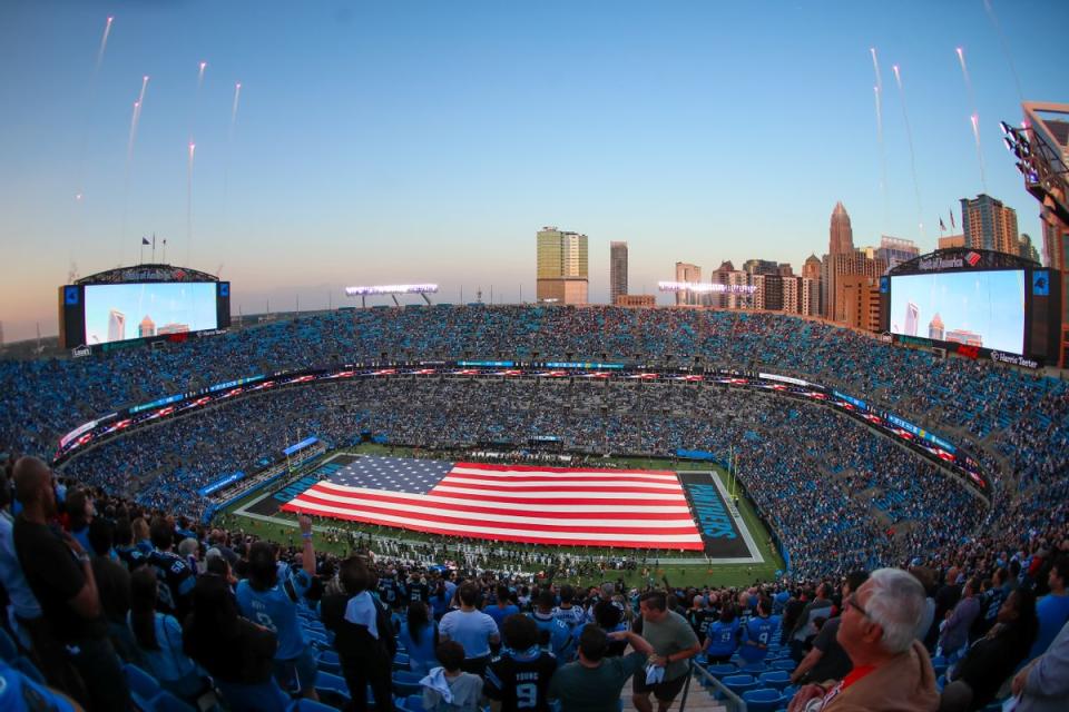 People surround a football field. On the field, a striped red-and-white flag with 50 white starts on a blue square in the corner of the flag covers the entire field. You can see fireworks surrounding the field. 