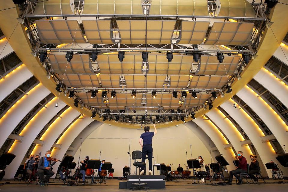 Musicians in masks are seated onstage as Gustavo Dudamel, in jeans, bounces on his toes and lifts a baton.