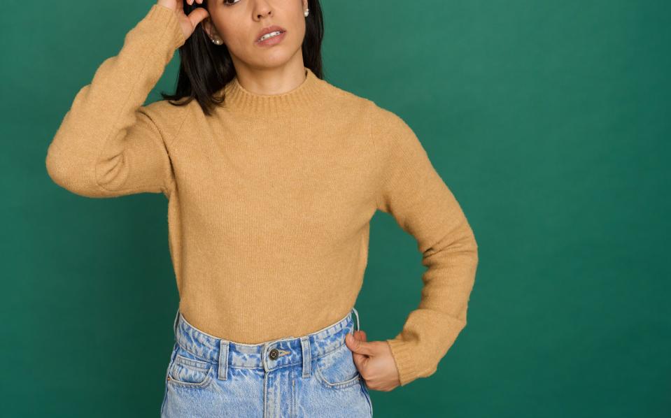 A woman with dark hair in a beige sweater and high-waisted jeans stands against a green background, scratching her head and looking puzzled