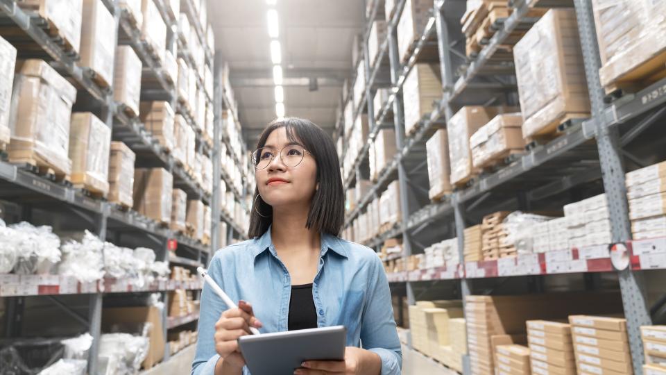 Candid of young attractive asian woman auditor or trainee staff work looking up stocktaking inventory in warehouse store by computer tablet with wide angle view.