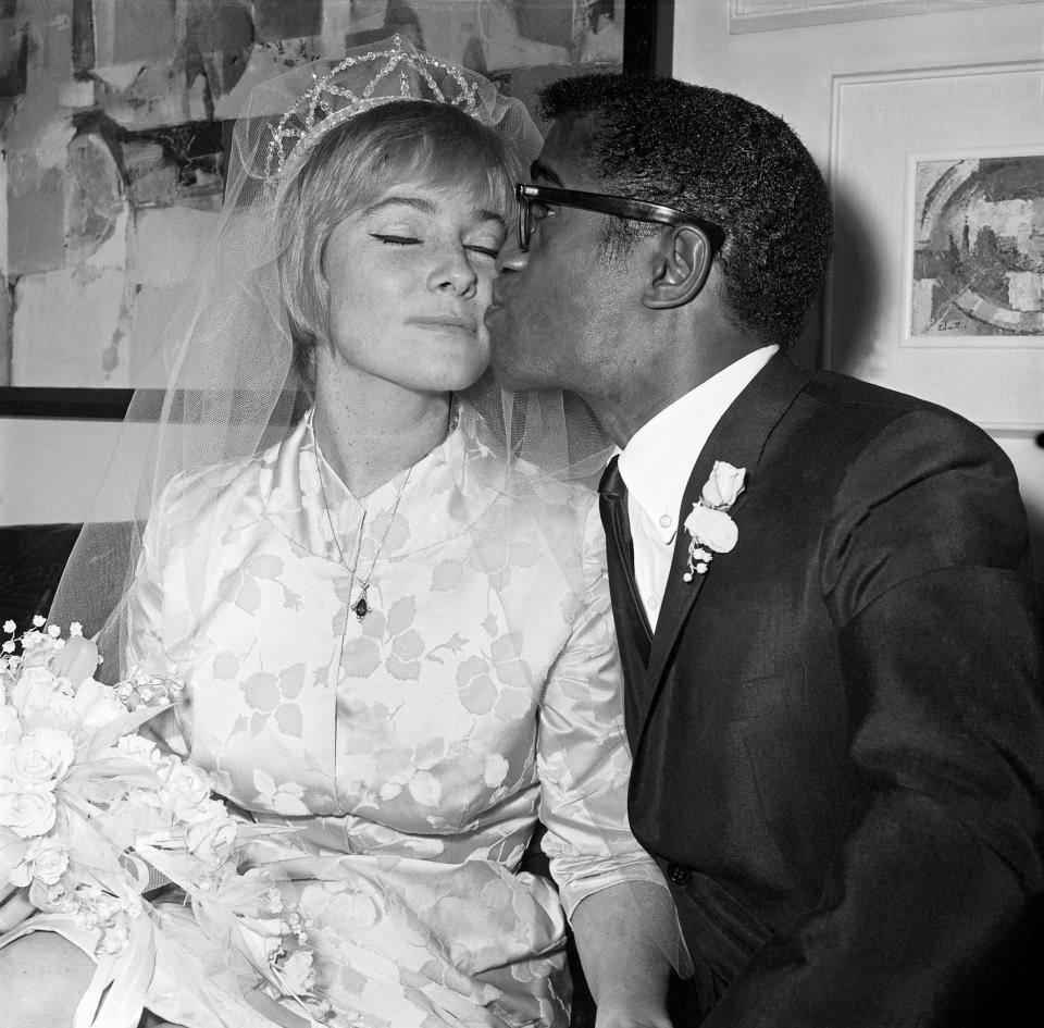 Sammy Davis Jr. is in a tux and he kisses May Britt on the cheek.