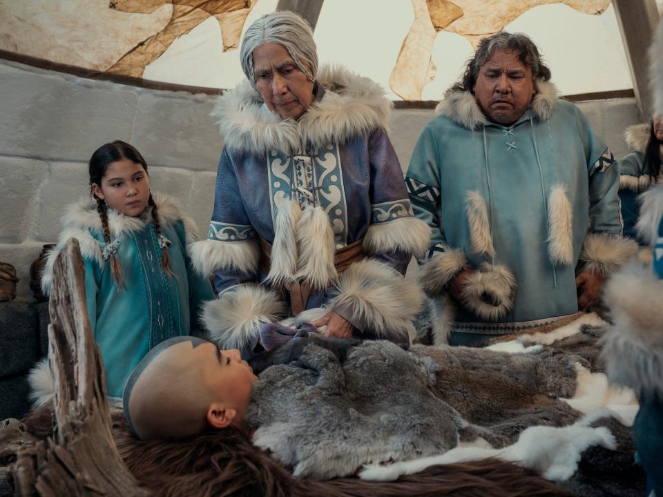 katara's gran gran in the avatar live action, a young girl, and a man in blue robes all stand over a sleeping aang, covered in furs