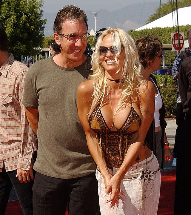 Tim Allen in an olive green t-shirt smiles behind Pamela Anderson in a low-cut tank top and white pants on the 2022 Teen Choice Awards carpet