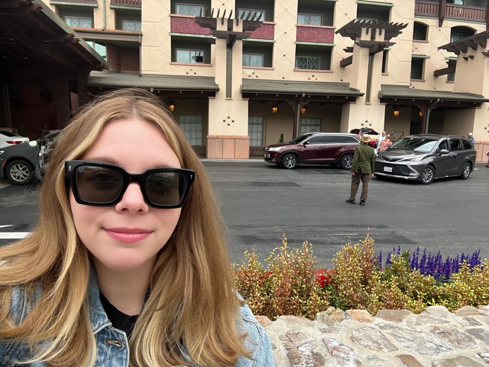 Jacqueline, wearing black sunglasses and a denim jacket, takes a selfie in front of Disney's Grand Californian Resort.