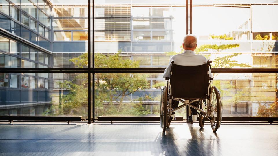 Senior Man in Wheelchair looking out of a window in a hospital corridor.