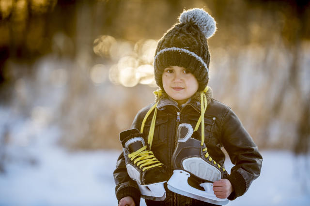 Young child standing outside in the winter with a pair of ice skate hanging around his neck, blurred background
