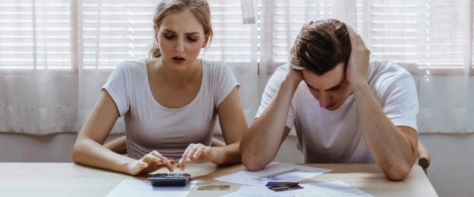 Stressed couple look at pile of bills, use a calculator, sitting at kitchen table