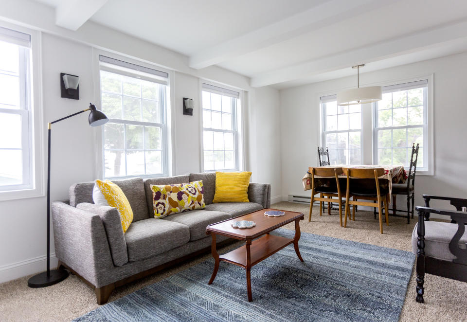 Modern living room with grey sofa, colorful pillows, blue rug, wooden coffee table, dining table with chairs, and windows with natural light