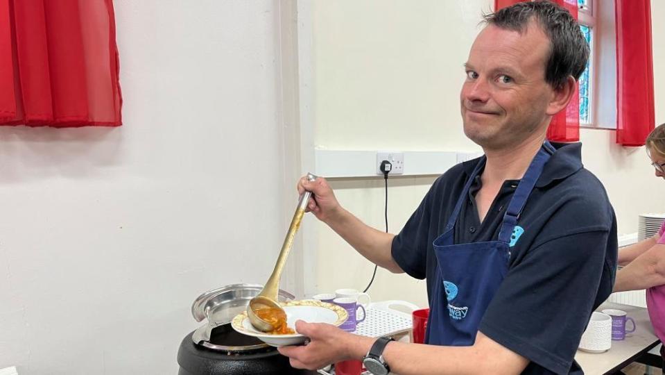 Paul Hubbard with short hair and wearing a blue apron ladles soup