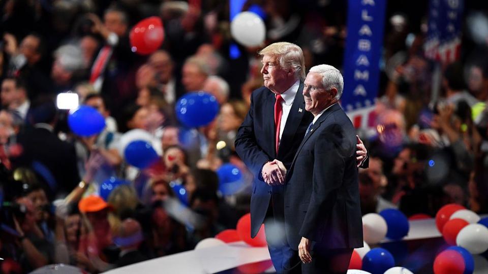 a profile shot of donald trump and mike pence standing on a stage shaking hands, both men wear suits, on the stage floor around them are red, white, and blue balloons and behind them is a blurred crowd