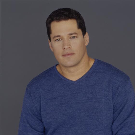 A young man in with dark hair in a blue sweater posing against a dark gray background