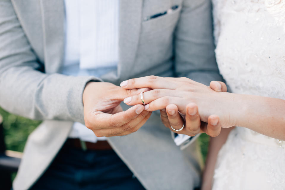 A close-up of a bride and groom exchanging rings. The groom is in a suit, and the bride wears a wedding dress with lace details. Names unknown