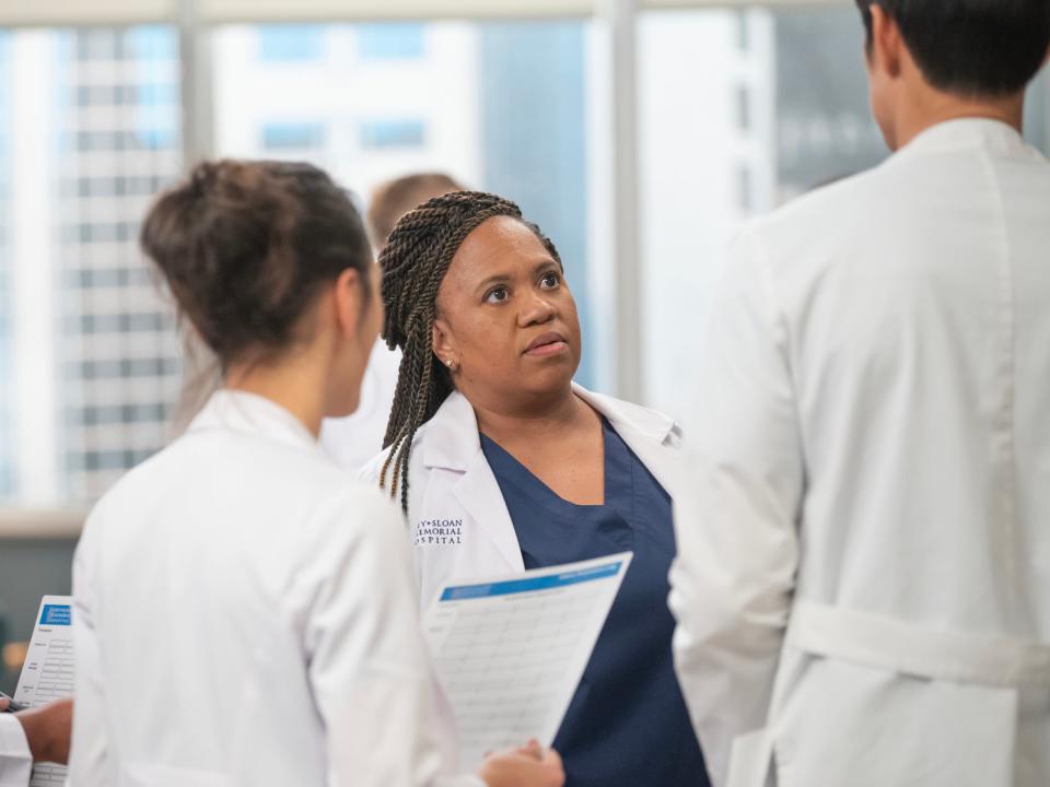 chandra wilson as miranda bailey in grey's anatomy. she has long braids pulled half back, dark blue scrubs, and a white doctor's coat. she's speaking to three others in white coats