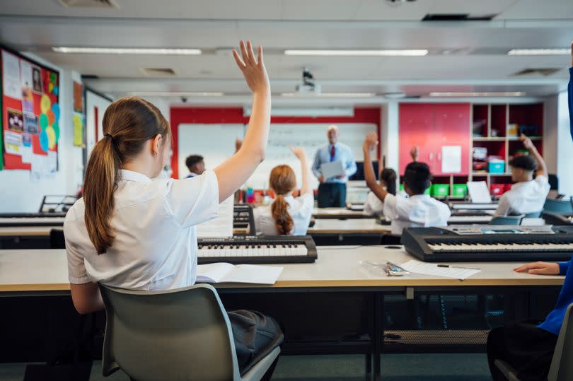 A wide-view shot of teenage music students in a classroom learning how to play the keyboard with their teacher at a school. The students are wearing school uniforms and the teacher is smartly dressed.