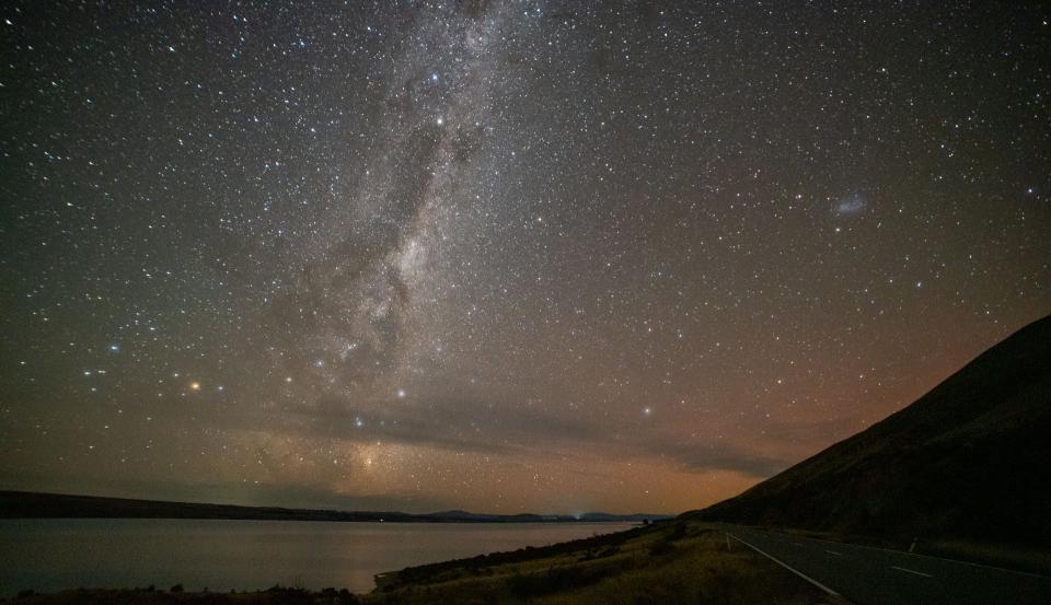 Photo of a road next to a lake with the night sky in the background clearly featuring the dark and light-grey band of the Milky Way Galaxy.