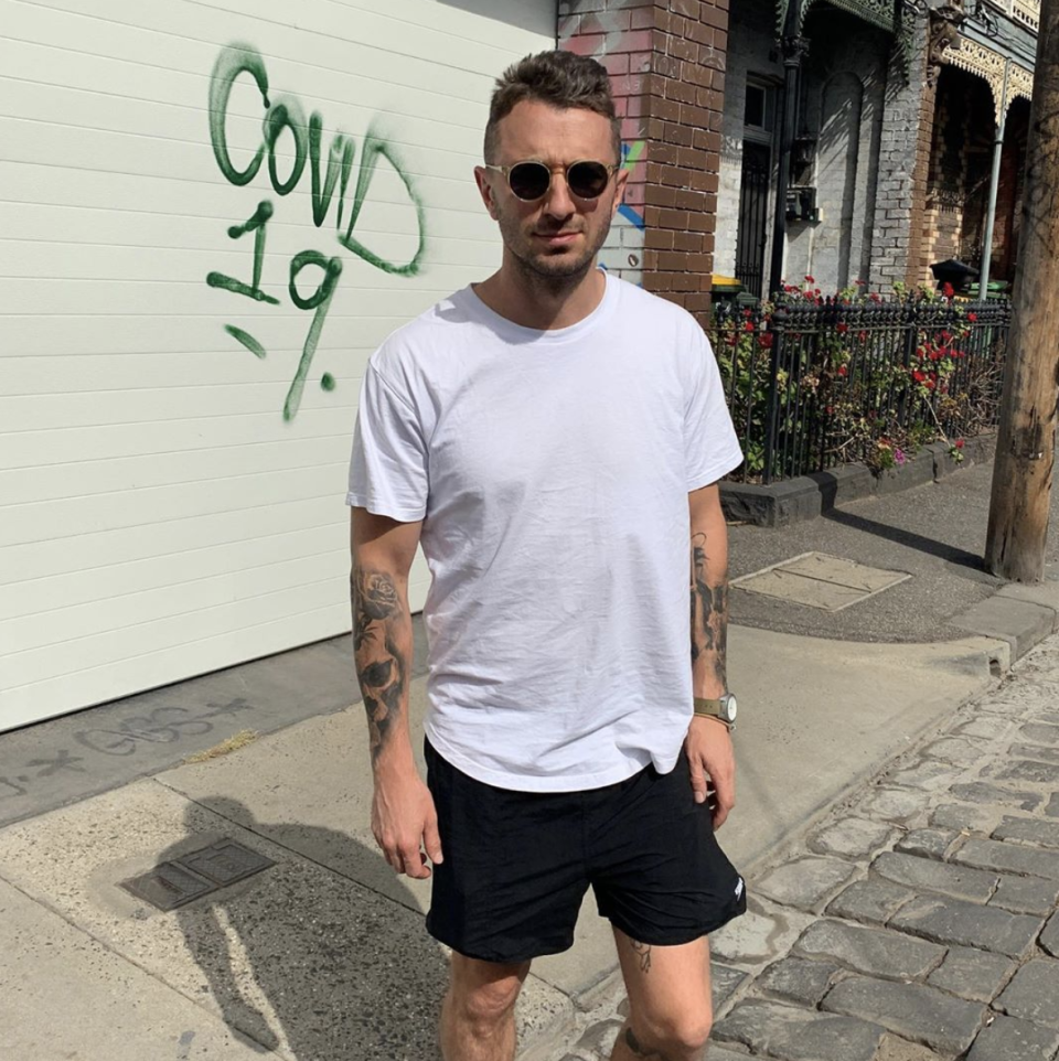 Australian comedian Tommy Little wearing a white t-shirt, black shorts and sunglasses standing on a street