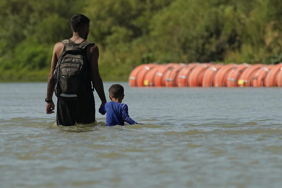 FILE - Migrants walk past large buoys being used as a floating border barrier on the Rio Grande, Aug. 1, 2023, in Eagle Pass, Texas. Texas must move a floating barrier on the Rio Grande that drew backlash from Mexico, a federal appeals court ruled Friday, Dec. 1, 2023, dealing a blow to one of Republican Gov. Greg Abbott's aggressive measures aimed at stopping migrants from entering the U.S. illegally. (AP Photo/Eric Gay, file)