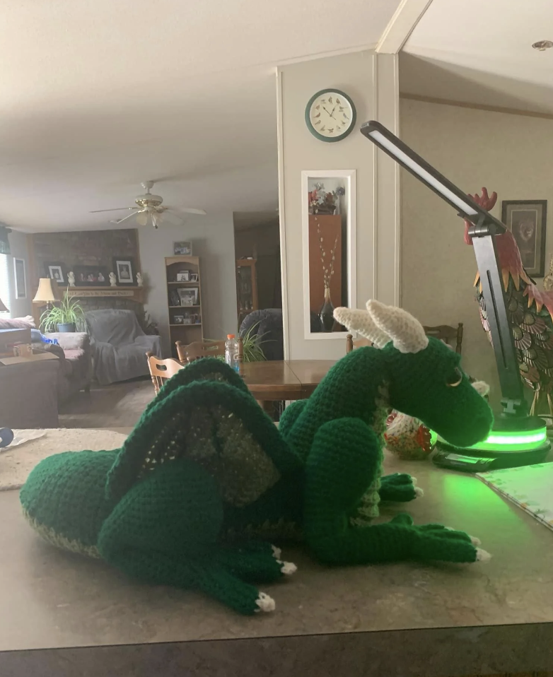 A handmade crocheted dragon is lying on a countertop in a cozy living room with various decorations and a clock on the wall