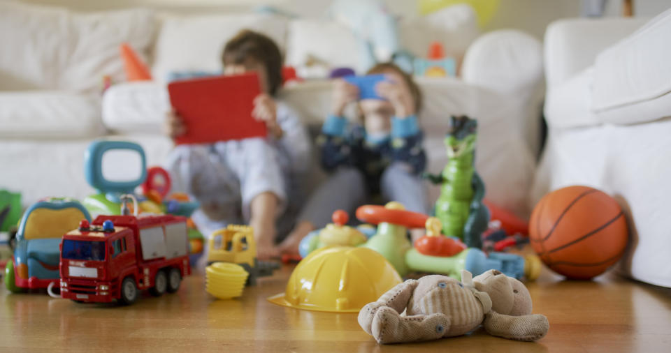 Two children sitting on a couch behind a variety of toys scattered on the floor, including a fire truck, construction hat, basketball, and teddy bear