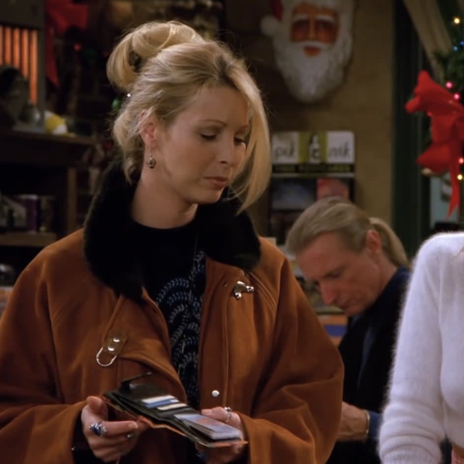 Lisa Kudrow as Phoebe Buffay, in a brown coat with black fur collar, holding a wallet in a cafe. Background includes Gunther and a Santa decoration